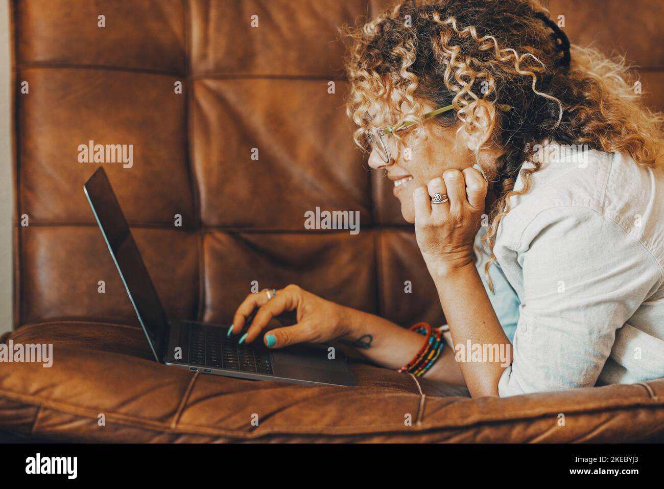Online-Suche im Web Konzept Menschen Aktivität. Glückliche Frau lächelt vor einem Computer, der auf dem Sofa liegt. Eine weibliche Person, die ihren Laptop für Freizeit oder Beruf benutzt, legt sich bequem auf die Couch Stockfoto