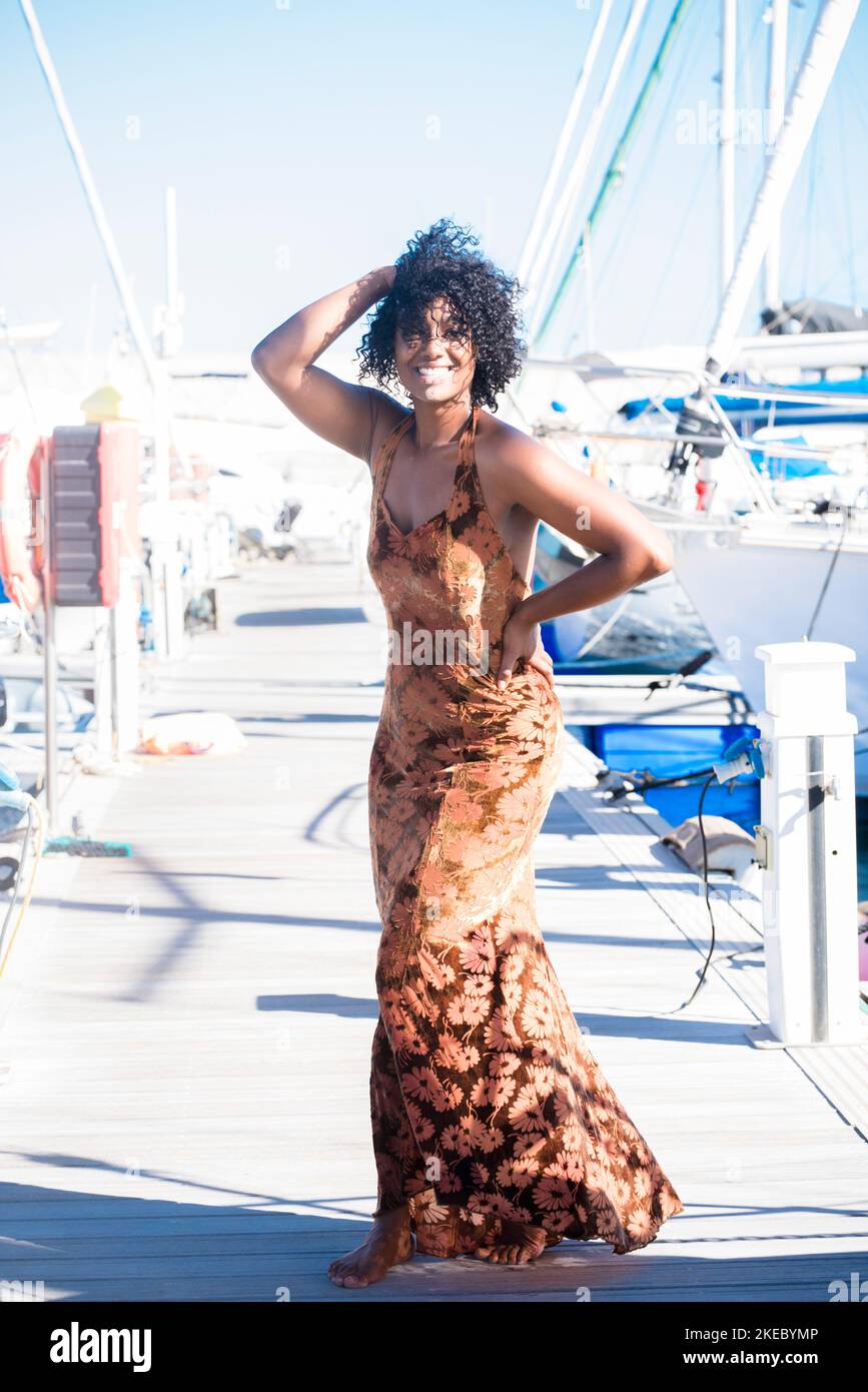 Porträt der schwarzen jungen Frau in elegantem Kleid in touristischen Outdoor-Freizeitaktivitäten. Ethnische verschönert Mädchen genießen Sonne und Urlaub am Hafen mit blauem Himmel im Hintergrund Stockfoto