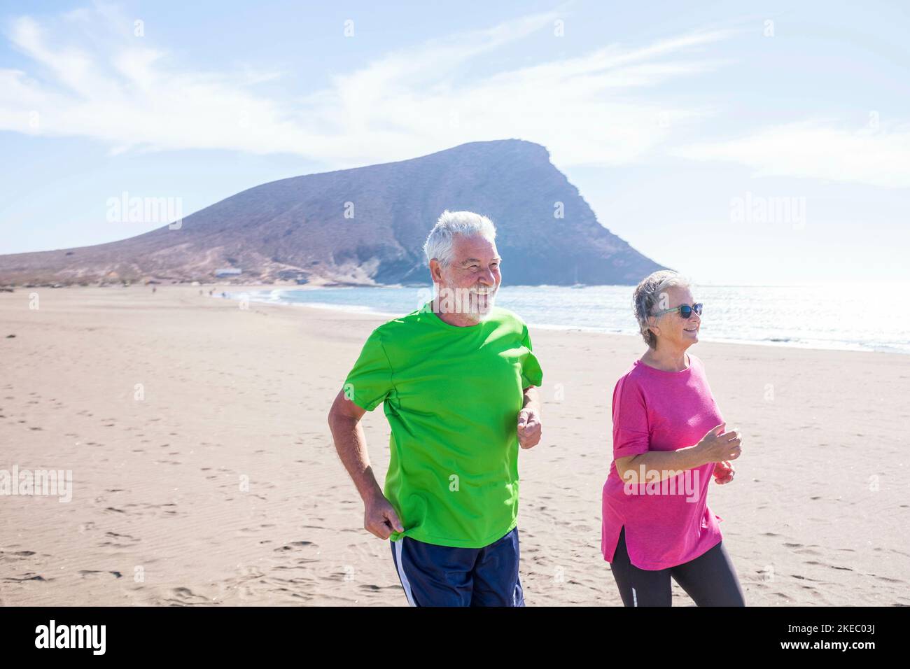 Ein paar von zwei reifen Menschen machen Bewegung und Sport zusammen Am Strand lächelnd und lachend - aktive Senioren tun Joggen und Laufen, um gesund und fit zu sein Stockfoto