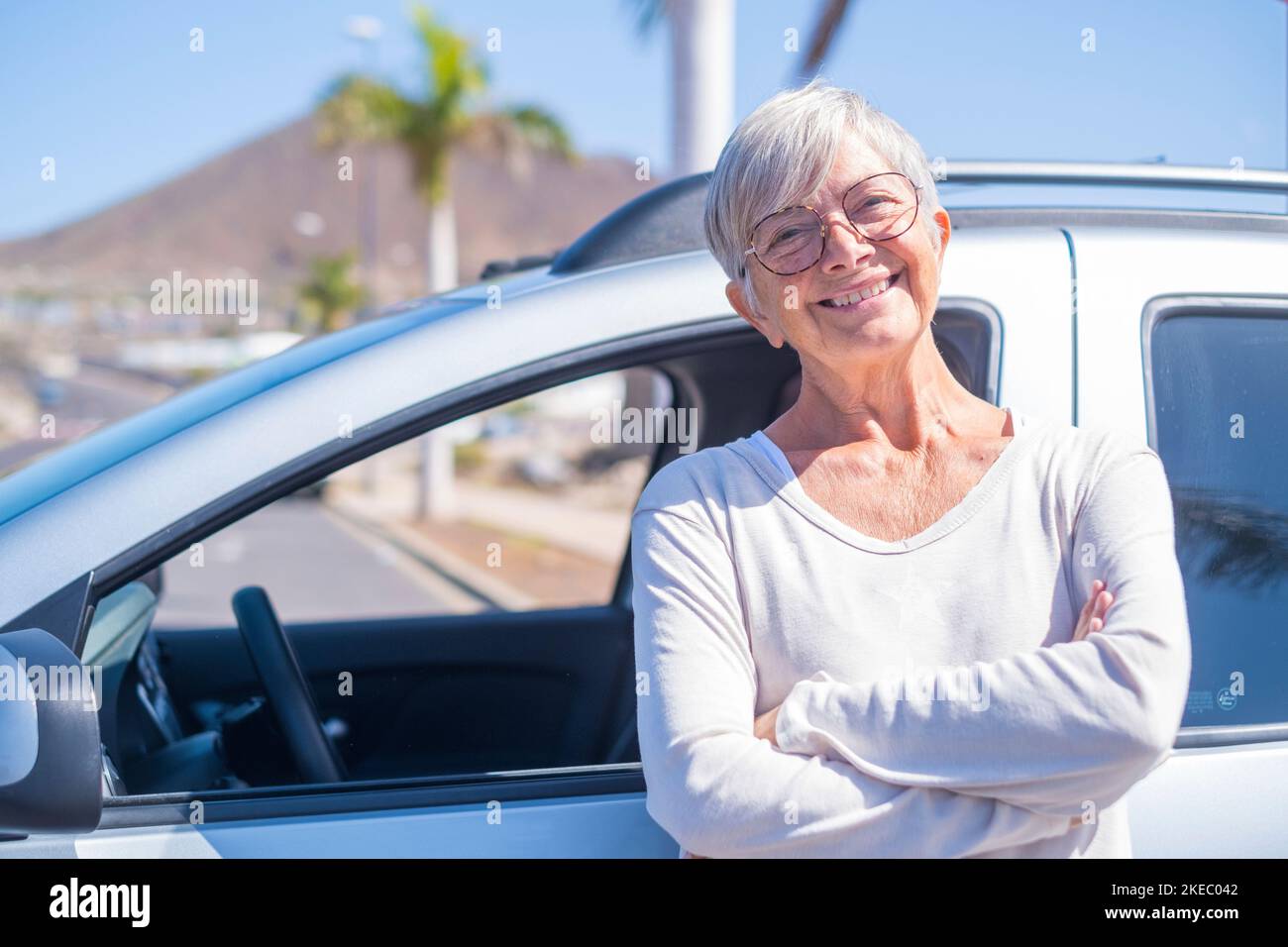 Nahaufnahme einer alten Frau, die lächelnd auf die Kamera blickt und ein Auto im Rücken hat. Porträt einer kaukasischen Frau, die neues Auto genießt. Zufriedener und zufriedener Kunde Stockfoto