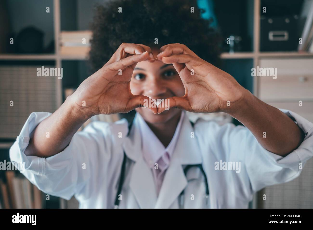 Frau tut Herzform Geste mit den Händen. Lächelnde schwarze Ärztin, die mit ihren Händen ein Liebessymbol macht. Mitarbeiter im Gesundheitswesen, der den Patienten Liebe und Unterstützung gibt Stockfoto
