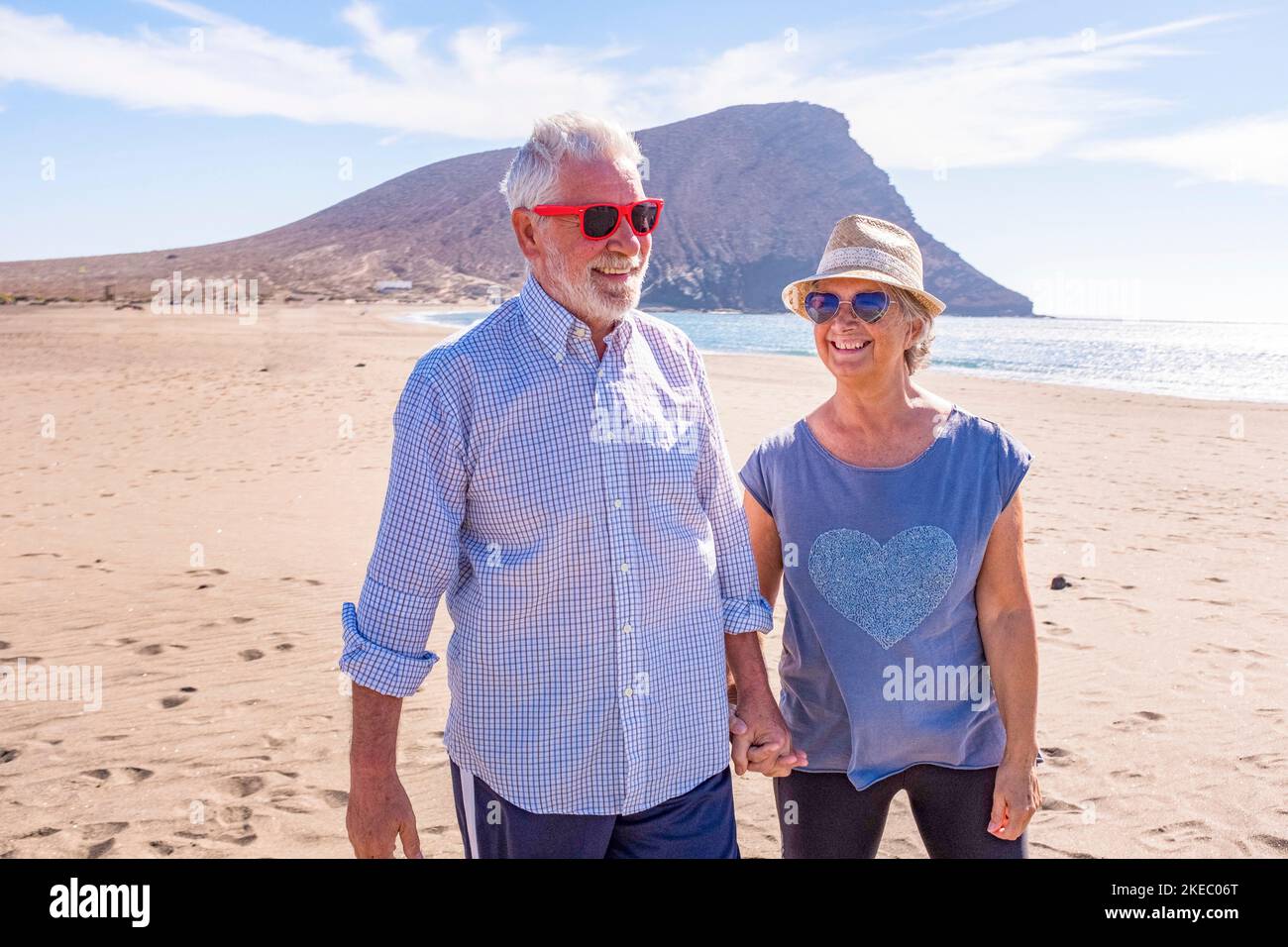 Ein paar ältere Leute, die Spaß haben, gemeinsam auf dem Sand des Strandes zu spazieren und ihren Urlaub zu genießen oder mit dem Meer oder dem Meer im Hintergrund in die Natur zu reisen - zwei Senioren, die sich verliebt haben, Spaß zu haben Stockfoto