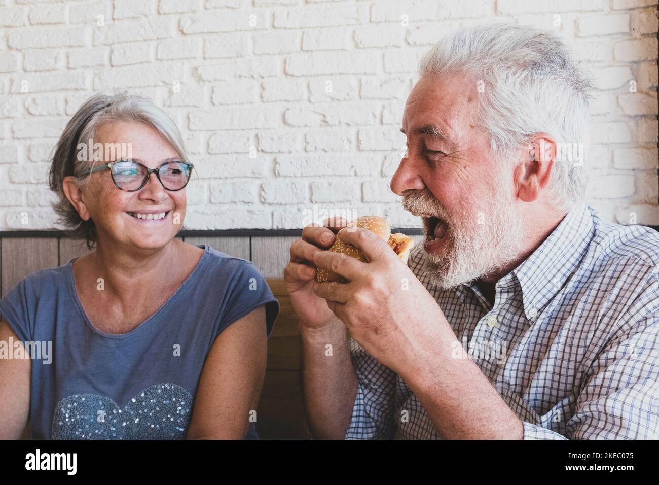 Ein paar Senioren zusammen essen und lachen - Reifer Mann hält einen Hamurger und isst ihn mit dem Mund Öffnete sich und seine Frau sah und lächelte Stockfoto