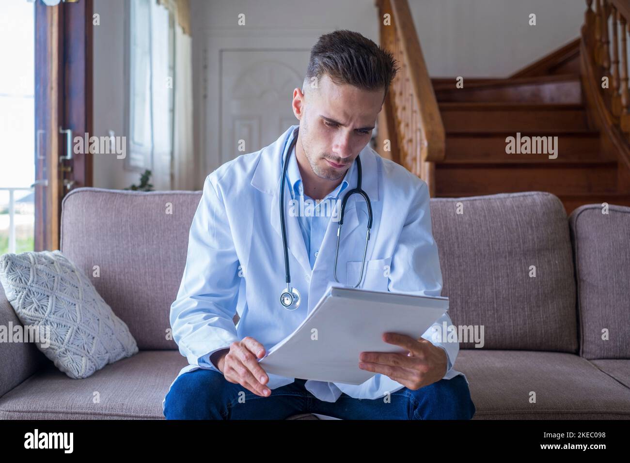 Porträt eines hübschen jungen männlichen Arztes in weißem Laborkittel mit Stethoskop, der Berichte im medizinischen Büro ansieht. Kaukasischer männlicher Gesundheitsarbeiter in Uniform, der schriftliche Berichte oder Testergebnisse des Patienten analysiert Stockfoto