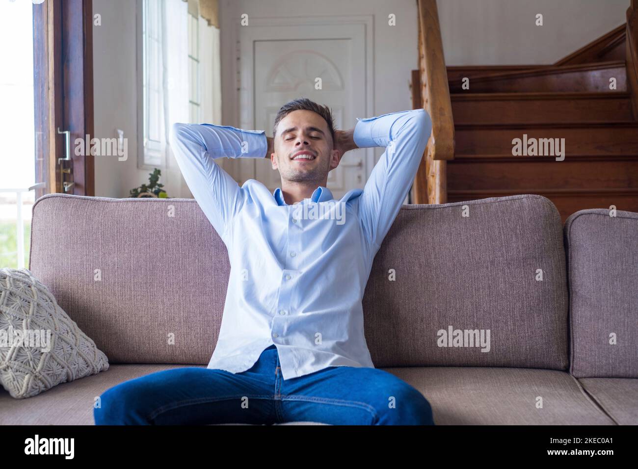 Zufriedener junger Mann, der sich zu Hause auf der Couch mit den Händen hinten entspannt. Lächelnder Mann mit geschlossenen Augen sitzt bequem auf der Couch im Wohnzimmer des modernen Hauses Stockfoto