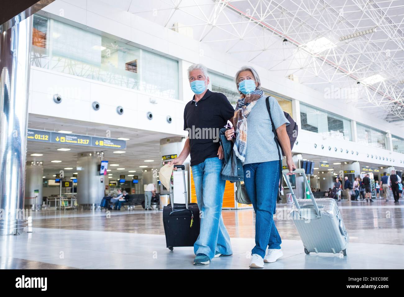 Zwei Senioren oder ältere Menschen, die am Flughafen zu ihrem Gate gehen und mit einer medizinischen Maske fliegen, um Viren wie Coronavirus oder Covid-19 zu verhindern - mit Gepäck oder Trolley Stockfoto