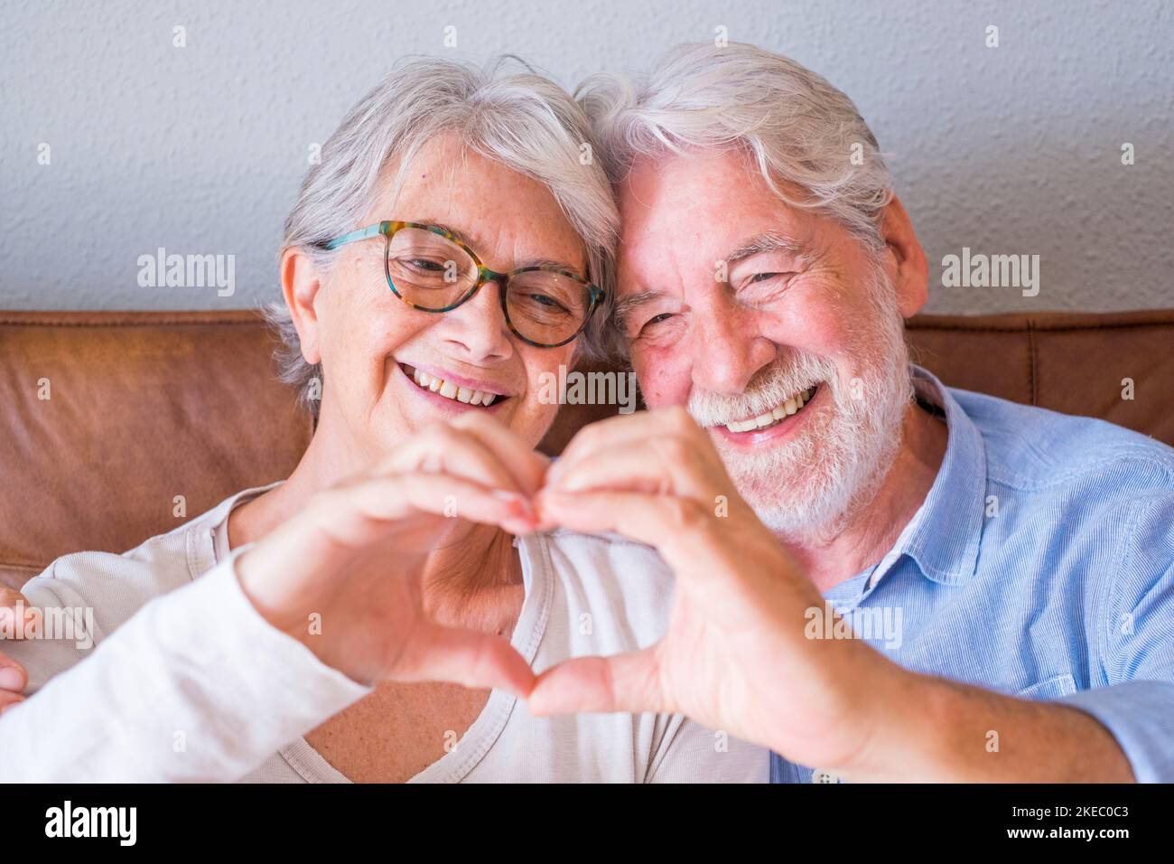 Porträt eines fröhlichen älteren Ehepaars, das sich umarmt und mit den Händen eine herzförmige Geste macht. Ältere glückliche Paar entspannen und posieren vor der Kamera sitzen im Wohnzimmer. Stockfoto
