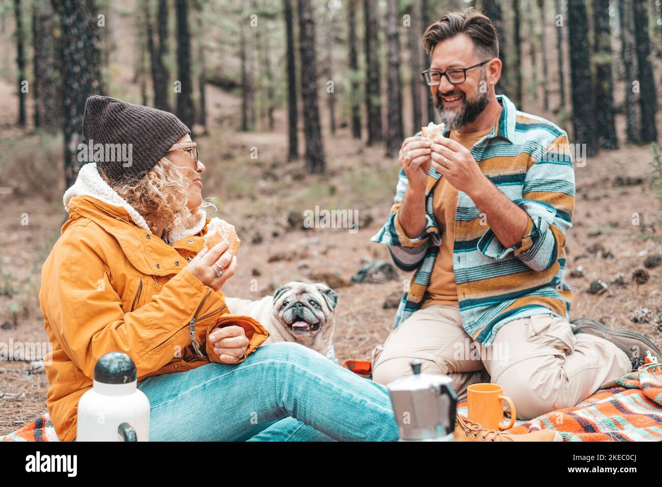 Zwei glückliche Menschen und ein Hund essen zusammen und machen ein Picknick auf dem Boden in den Bergen mit vielen Bäumen um sie herum Stockfoto