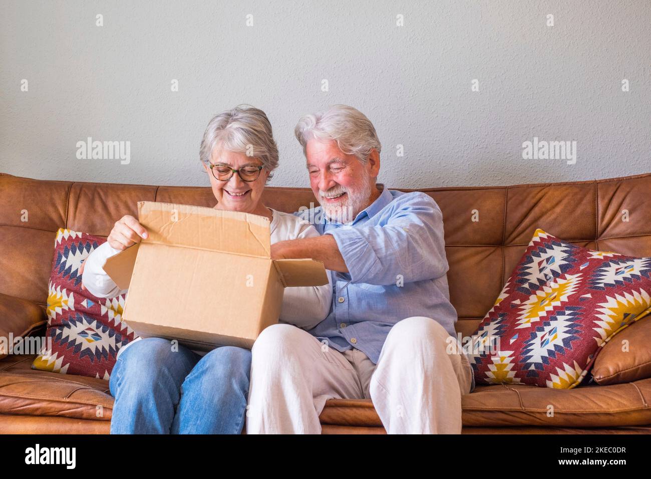 Altes Paar, das zu Hause den Lieferkarton auspackte. Glückliches Seniorehepaar, das im Wohnzimmer zu Hause auf dem Sofa saß und die Kartonschachtel ansah. Ein älteres Paar, das auscheckte, brachte Sachen zu Hause. Stockfoto