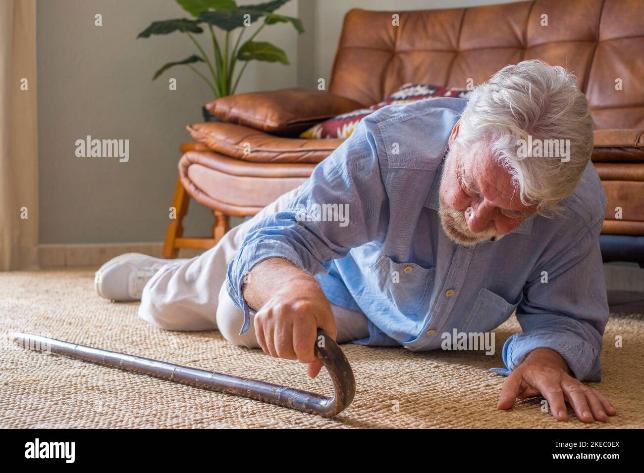 Älterer Mann, der auf dem Boden lag, nachdem er mit einem hölzernen Spazierstock neben der Couch auf dem Teppich im Wohnzimmer zu Hause heruntergefallen war. Alter Mann, der unter Schmerzen leidet und nach einem Sturz zu Hause um aufzustehen kämpft Stockfoto
