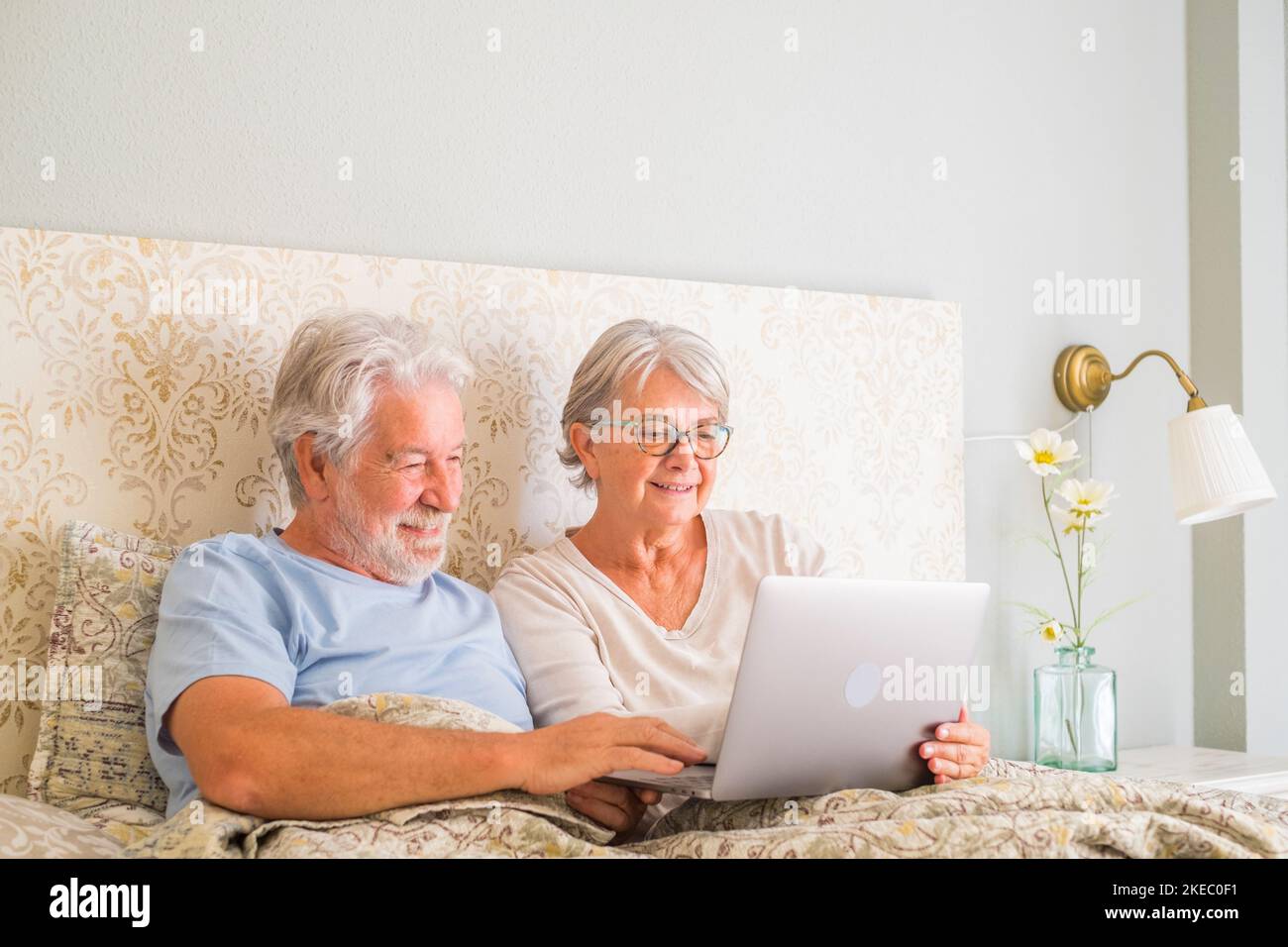 Lächelndes Seniorenpaar, das zu Hause mit einem Laptop im Bett Social Media-Inhalte durchsucht und ansieht. Entspannter alter Mann und Frau verbringen Freizeit im Bett, teilen Online-Film im Schlafzimmer in einem modernen Apartment Stockfoto