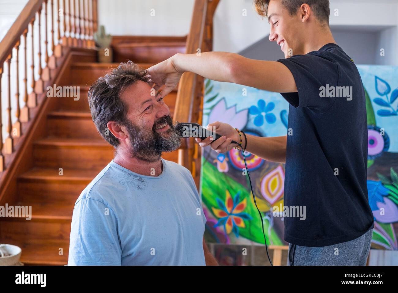 Ein Paar von Vater und Sohn zu Hause lachen und Spaß haben zusammen den Bart des Vaters in Quarantäne schneiden - lächeln und spielen auf Be a Coiffeur oder Friseur hausgemacht Stockfoto