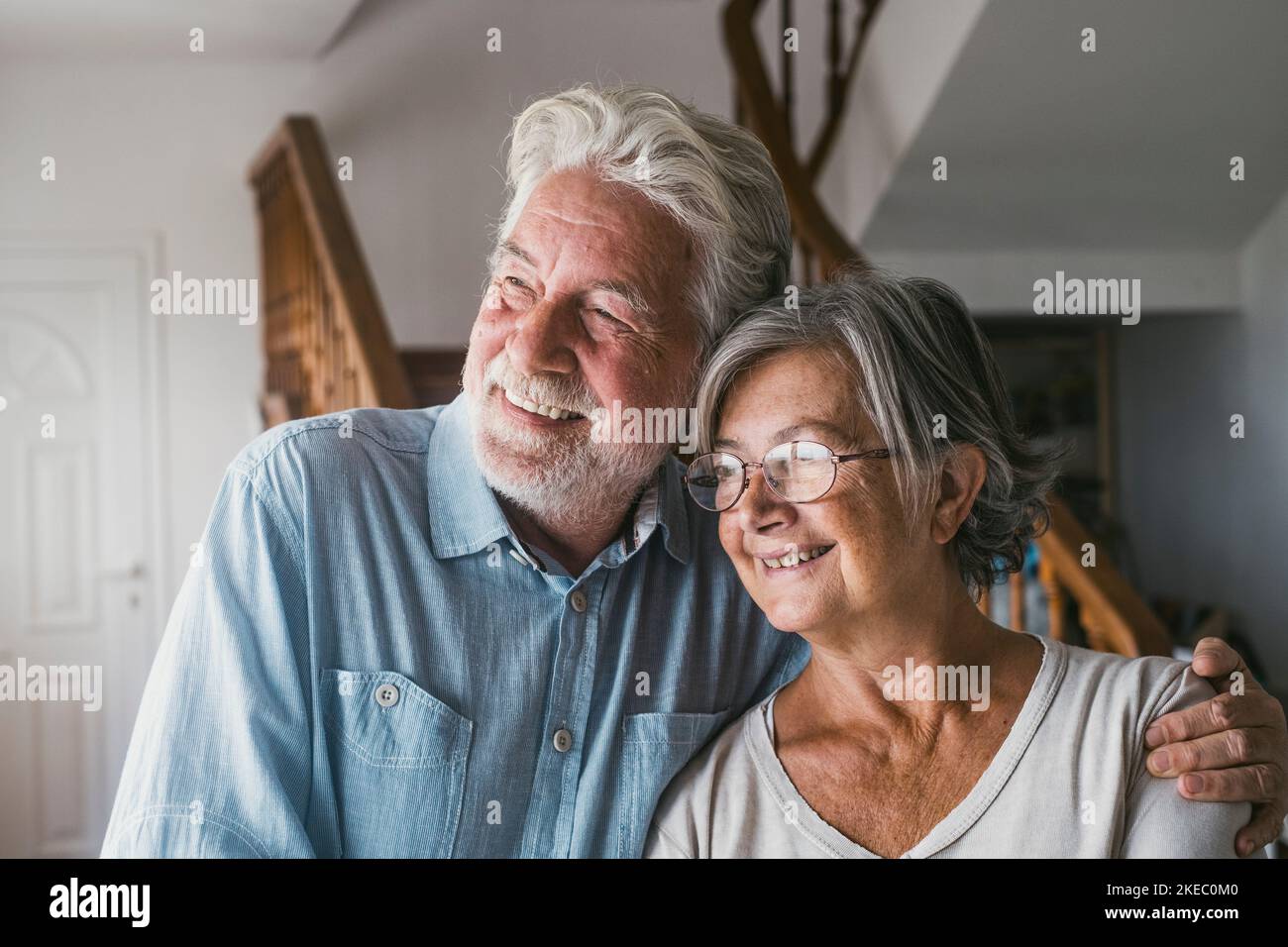 Ein Paar, das sich an einem hellen, sonnigen Tag zu Hause entspannt und durch das Fenster blickt. Glücklicher alter Mann und Frau blicken weg und bewundern die Aussicht vom modernen Haus. Lächelndes, älteres Paar zu Hause Stockfoto