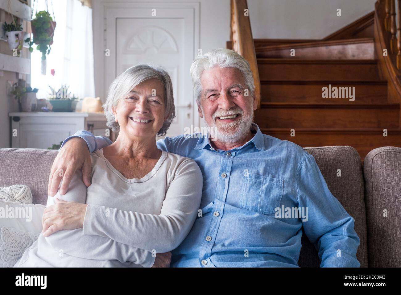 Porträt eines glücklichen Rentnerpaares mittleren Alters, das sich zu Hause auf dem gemütlichen Sofa entspannt. Lächelnd aufrichtig liebende ältere Hausbesitzer, die Kamera betrachten, für Fotos posieren, Liebe und Fürsorge in Innenräumen zeigen. Stockfoto