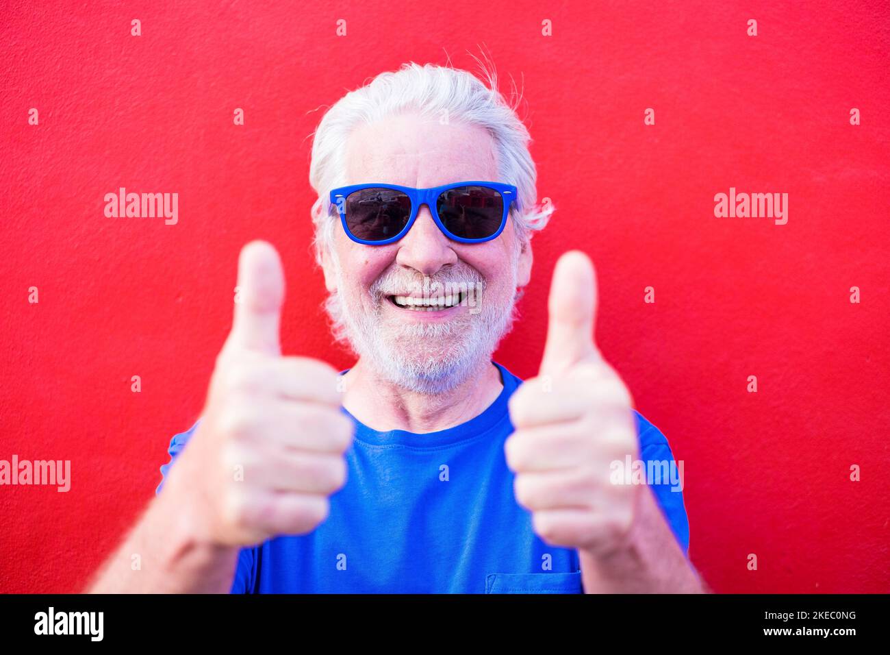 Porträt und Nahaufnahme von reifen und alten Mann trägt Blaue Sonnenbrille und lächelnd Blick auf die Kamera - Fröhlich und glücklich Senior mit rotem Hintergrund genießen und haben Spaß Stockfoto