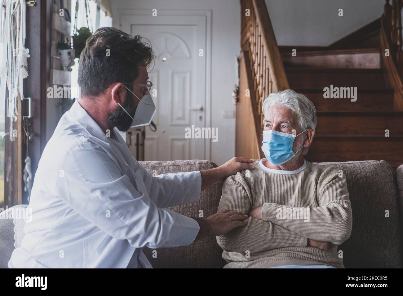 Männliche professionelle Arzt Beratung Senior Patient während der medizinischen Versorgung Besuch trägt Masken. Ein junger Arzt und ein alter älterer Herr, der auf dem Sofa sitzt und medizinische Hilfe leistet. Ältere Menschen in der häuslichen Pflege. Covid- und Coronavirus-positiver Test Stockfoto