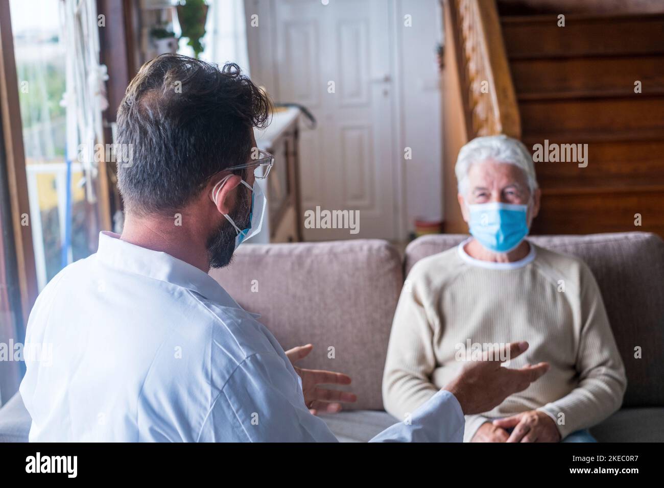 Männliche professionelle Arzt Beratung Senior Patient während der medizinischen Versorgung Besuch trägt Masken. Ein junger Arzt und ein alter älterer Herr, der auf dem Sofa sitzt und medizinische Hilfe leistet. Ältere Menschen in der häuslichen Pflege. Covid- und Coronavirus-positiver Test Stockfoto