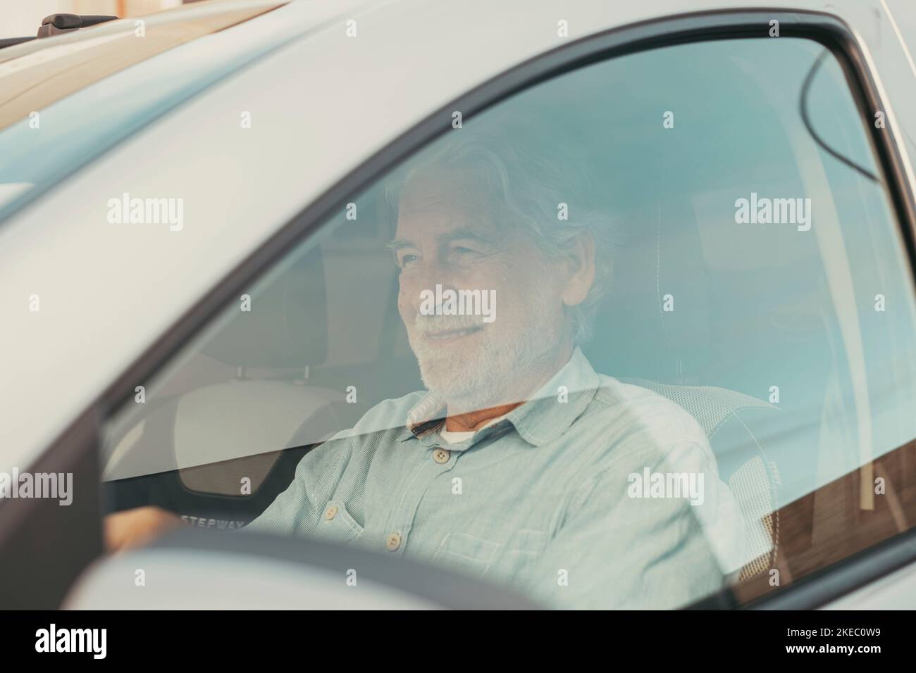 Glücklicher Besitzer. Gutaussehender bärtiger reifer Mann, der entspannt in seinem neu gekauften Auto sitzt und fröhlich aus dem Fenster blickt. Ein alter Senior fährt und hat Spaß. Stockfoto