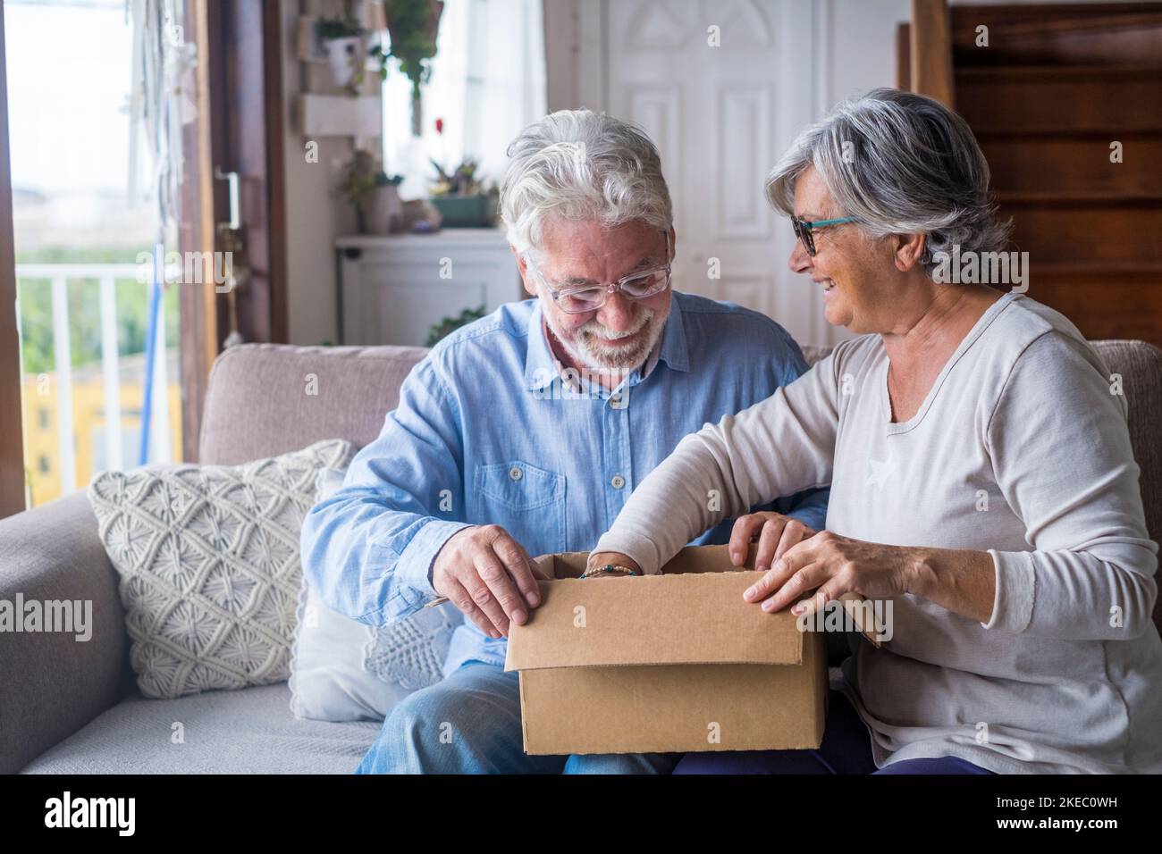 Altes Paar, das zu Hause den Lieferkarton auspackte. Glücklicher älterer Mann und Frau, die auf Karton schauten, während sie auf dem Sofa im Wohnzimmer saßen. Der ältere Mann und die ältere Frau, die auscheckten, brachten uns Sachen auf die Couch Stockfoto