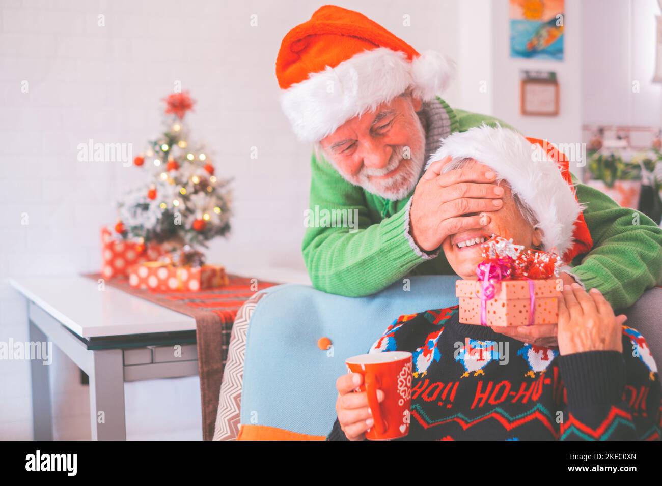 Seniorenpaar in warmer Kleidung und weihnachtsmütze, Mann, der die Augen der alten Frau mit einem Überraschungsgeschenk für sie bedeckt. Liebende alte romantische heterosexuelle Paar feiern weihnachtsfest zusammen Stockfoto