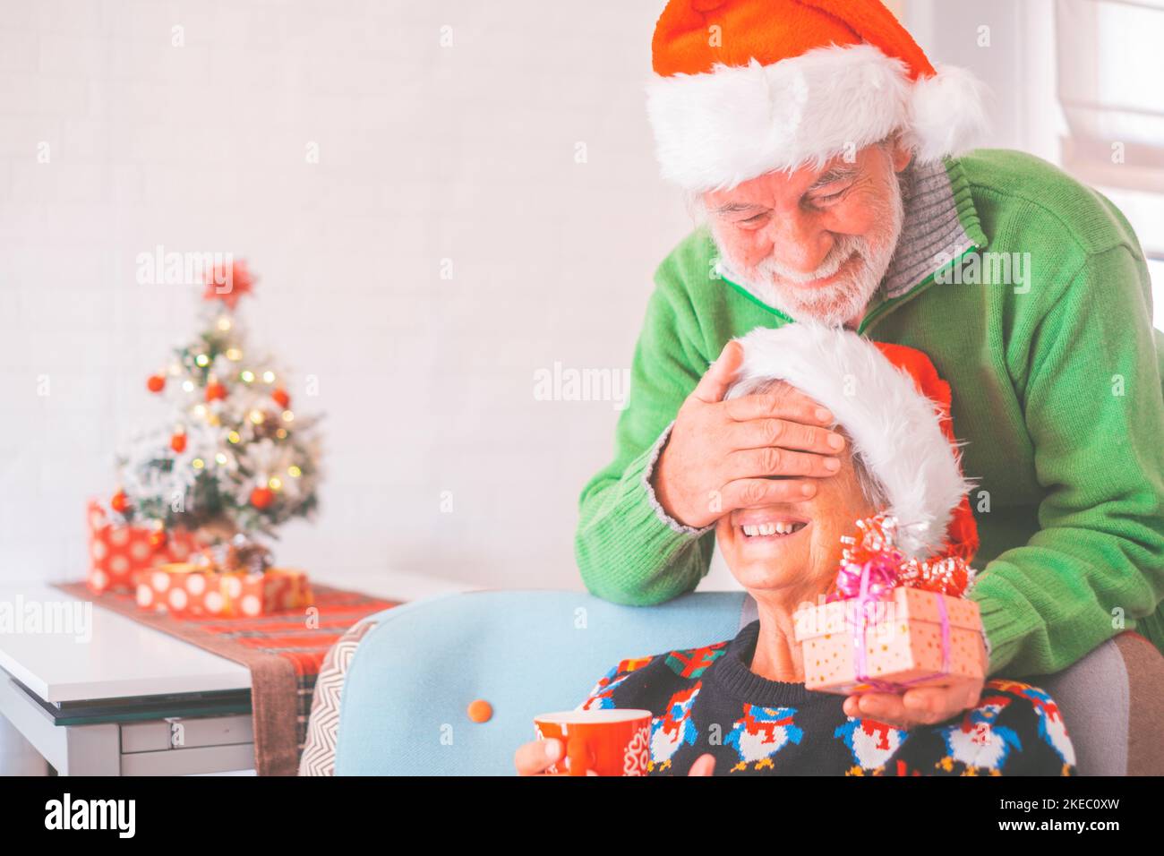 Seniorenpaar in warmer Kleidung und weihnachtsmütze, Mann, der die Augen der alten Frau mit einem Überraschungsgeschenk für sie bedeckt. Liebende alte romantische heterosexuelle Paar feiern weihnachtsfest zusammen Stockfoto