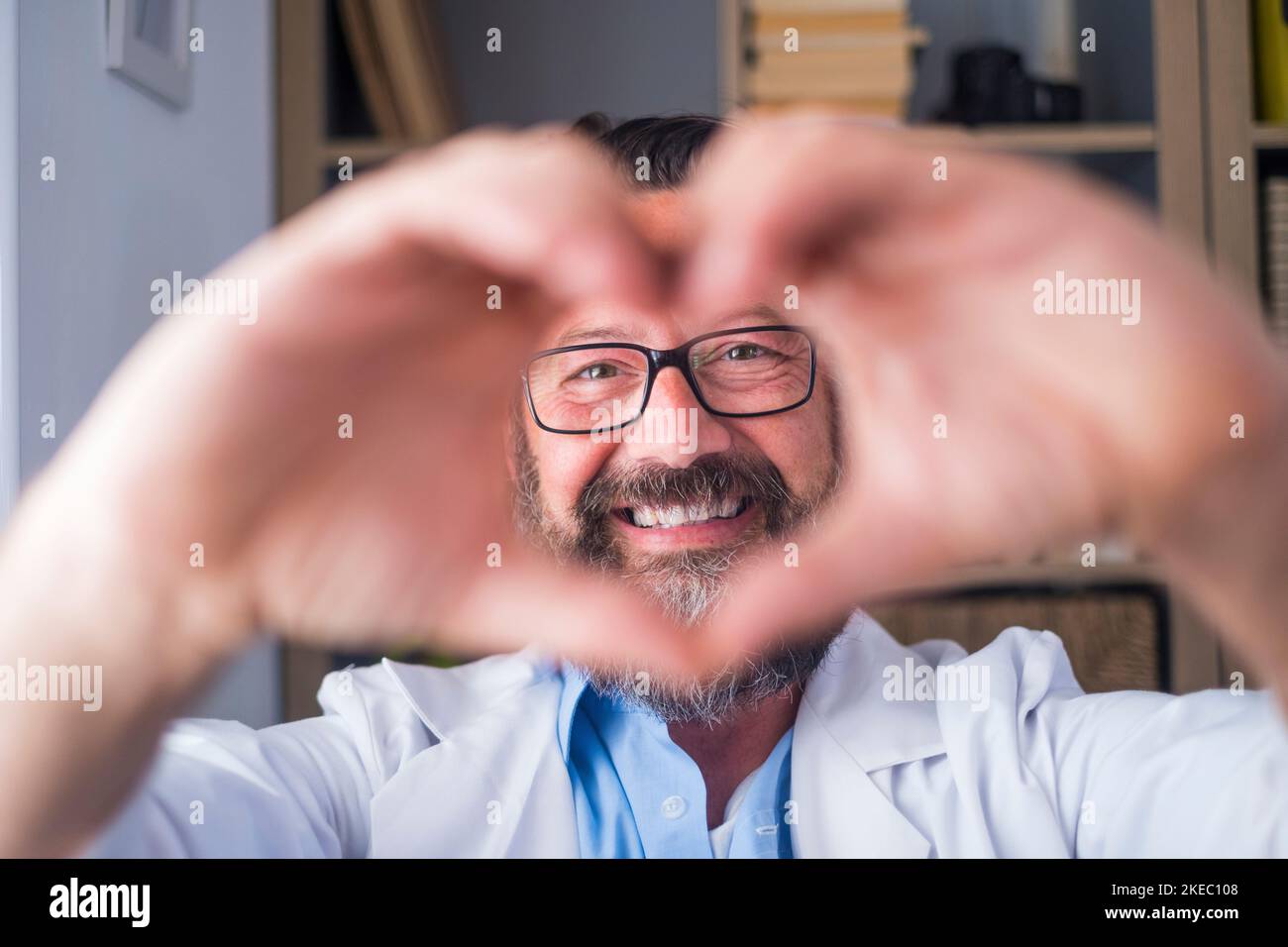 Mann macht Herz Form Geste mit den Händen. Lächelnder hübscher kaukasischer Arzt, der mit seinen Händen ein Liebessymbol macht. Mitarbeiter im Gesundheitswesen, der den Patienten Liebe und Unterstützung gibt Stockfoto