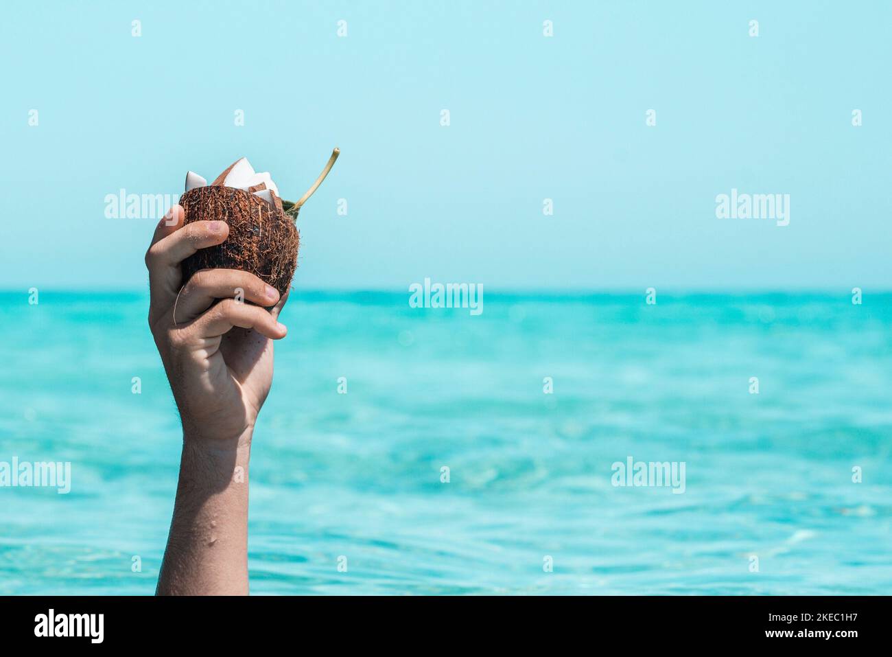 Nahaufnahme einer nicht erkennbaren Person, die eine halbe Kokosnussschale mit Scheiben auf Kokosnuss in sich gegen Meer und Himmel hält. Nasse Hand im Sommer mit Kokosnussschale. Erhobene Hand hält Kokosnuss gegen das Meer Stockfoto