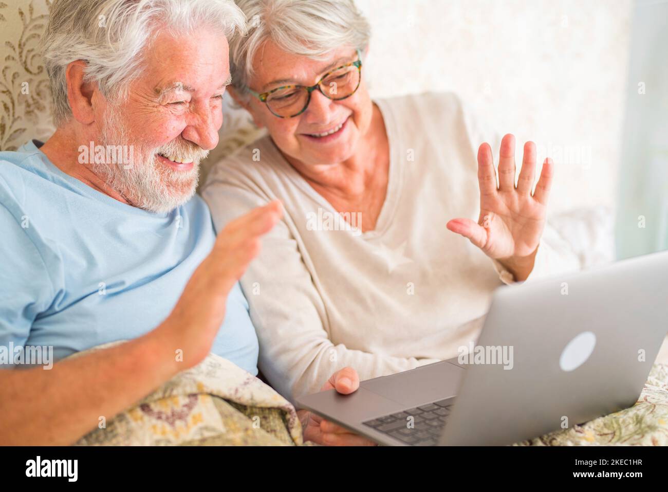 Altes, älteres kaukasisches Paar lächelt und chattet sich morgens mit einem Laptop im Schlafzimmer zu Hause im Bett. Ein älteres Paar begrüßt zu Hause einen Videoanruf auf dem Laptop. Stockfoto
