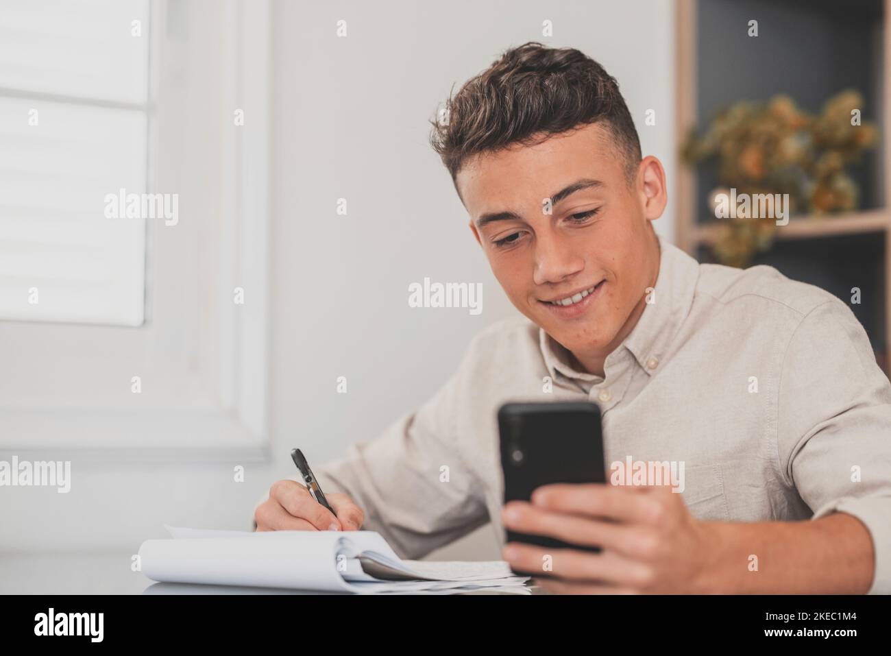Glücklicher junger Mann, der sein Smartphone zu Hause benutzt, um Hausaufgaben zu machen und alleine in der Halle Spaß zu haben. Männliche Teenager mit Telefon genießen Freizeit studieren und arbeiten. Stockfoto