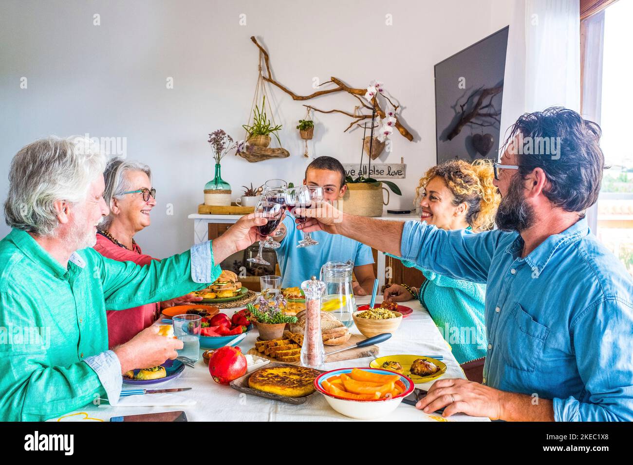 Tisch voller Lebensmittel zu Hause mit Menschen aller Alter essen zusammen und klirren mit Wein lächeln und haben Spaß - Familie, die drinnen zu Mittag essen Stockfoto