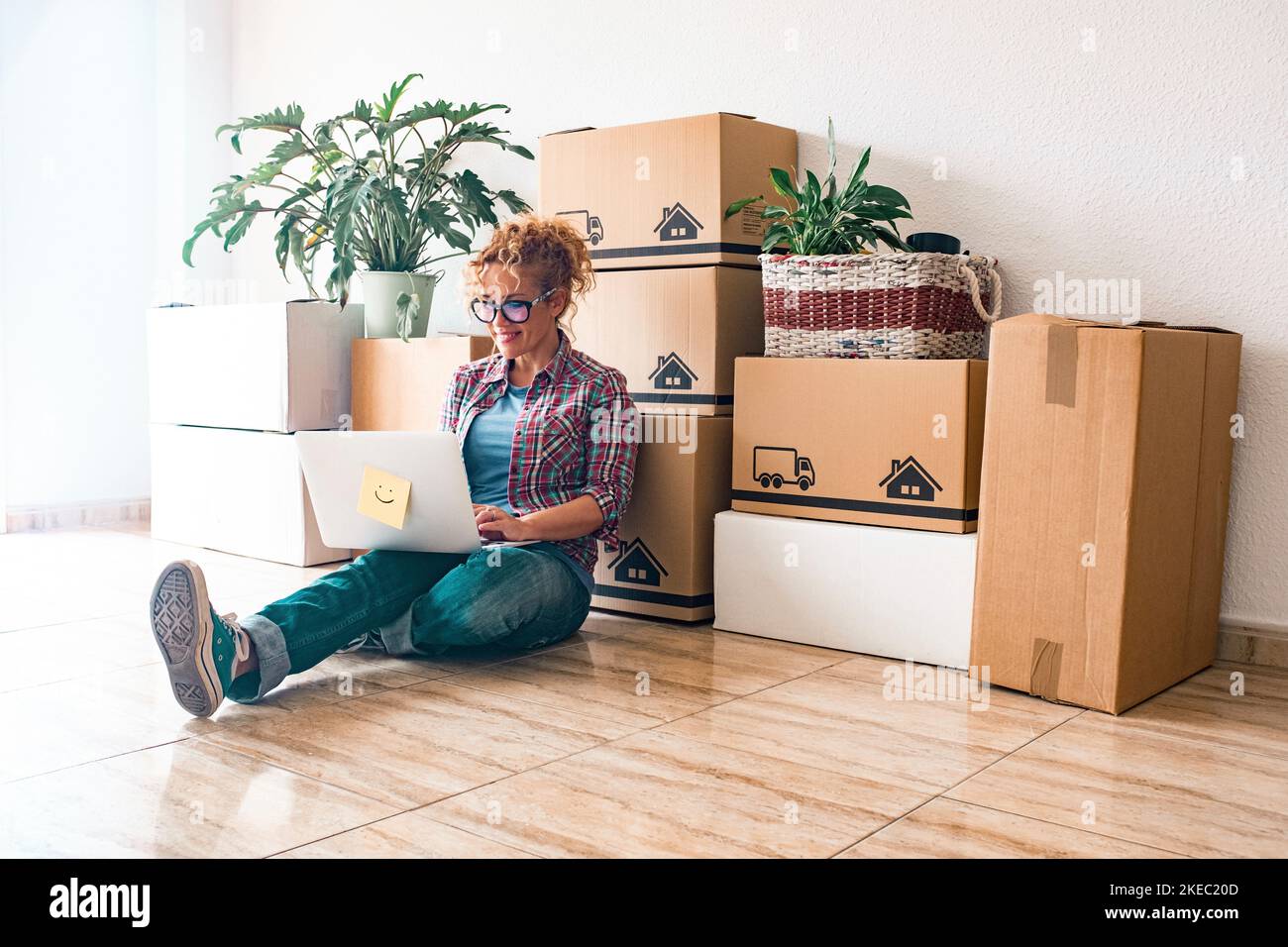 Eine junge Frau und glückliche Menschen kaufen ein neues Haus oder eine Wohnung zusammen, um zusammen zu leben - Person auf dem Boden mit Laptop mit Boxen und Packungen auf dem Rücken Stockfoto