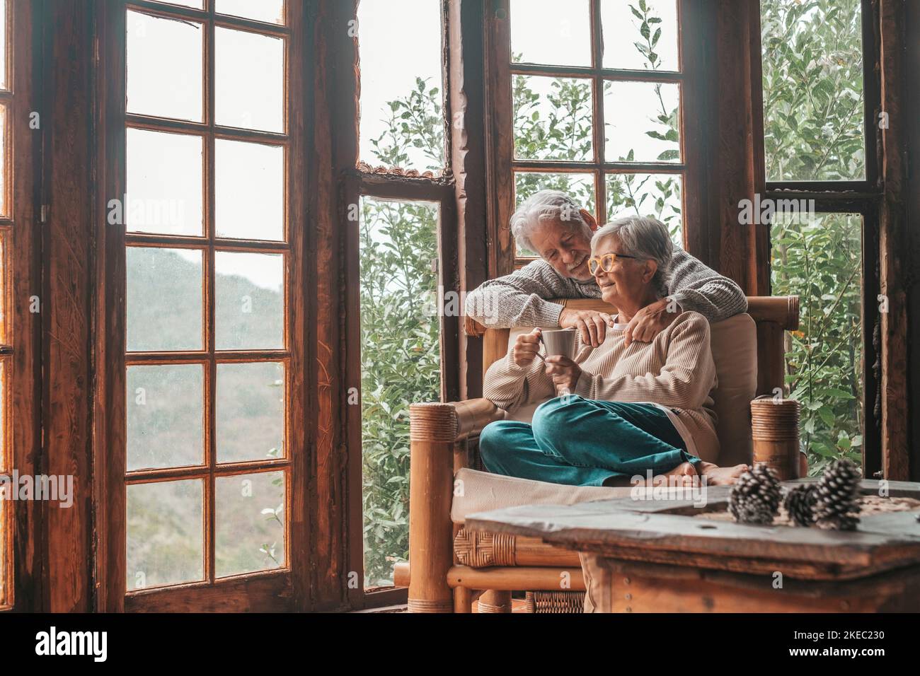 Ein glückliches, altes kaukasisches Paar, das Freizeit mit Blick durch das Fenster zu Hause verbringt. Liebevoller Ehemann umarmt Frau von hinten, sitzt auf einem Sessel im Haus und bewundert den Blick aus transparentem Türglas. Stockfoto