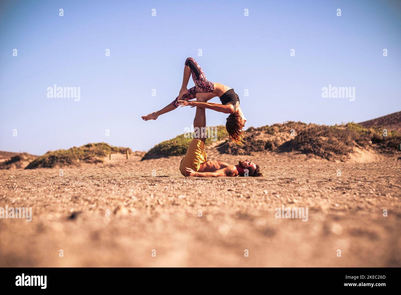 Zwei Erwachsene machen zusammen acroyoga am Strand Auf dem Sand - Mann hält seinen Teamkollegen mit seinem Beine Stockfoto