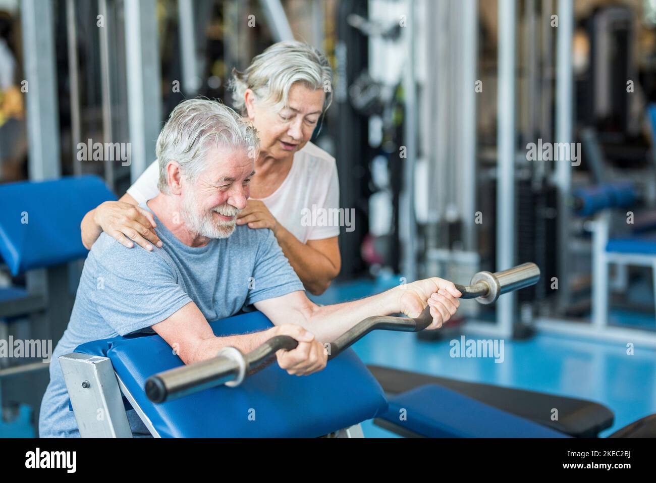 Ein paar von zwei Senioren im Fitnessstudio machen zusammen Sport Spaß haben, gesund und fit zu sein - Mann hält Ein barr ohne Gewicht und mit seiner Frau, die ihm hilft Stockfoto