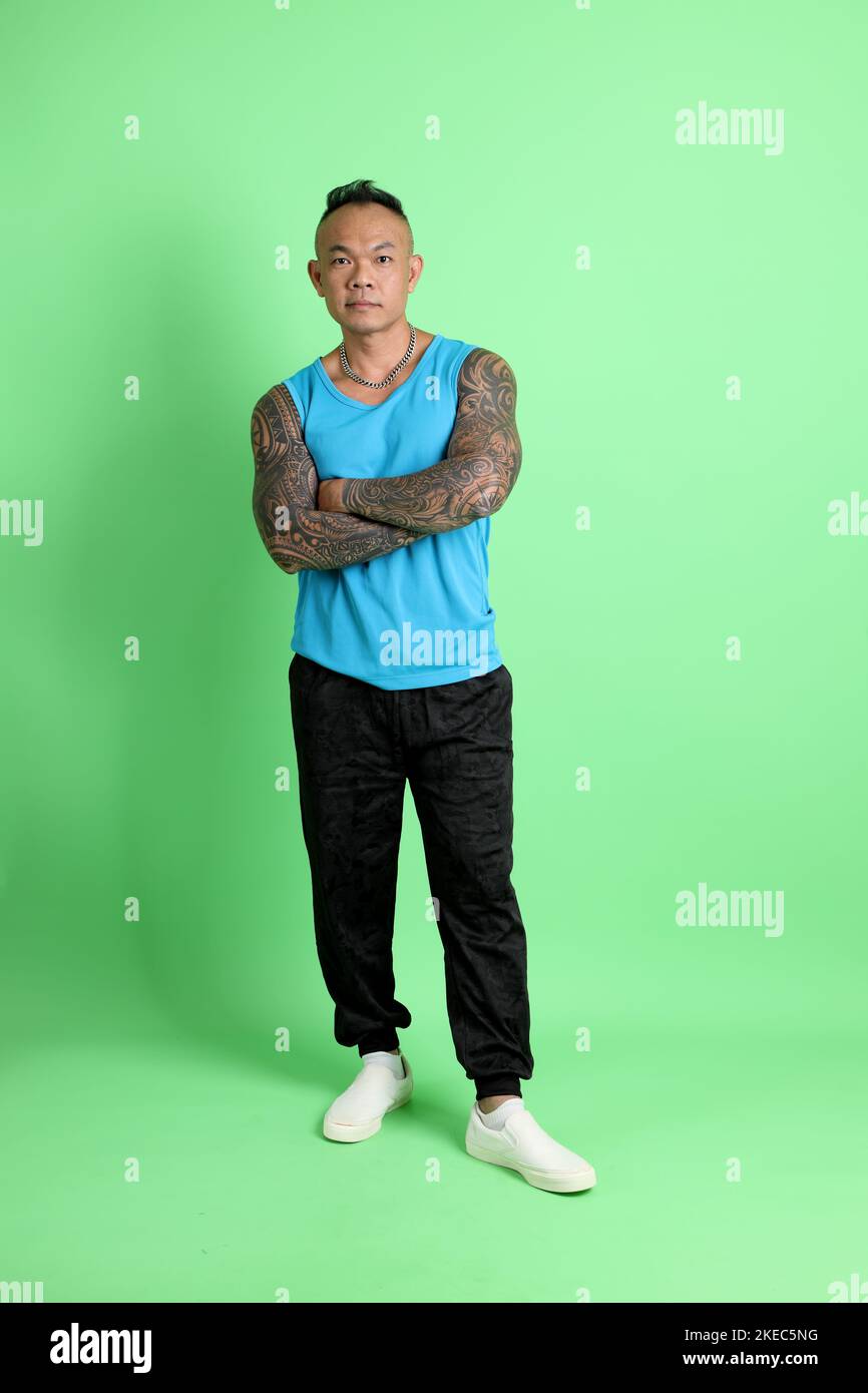 Der 40s-jährige asiatische Mann mit Tattoo, der auf dem grünen Hintergrund mit Sportkleidung steht. Stockfoto