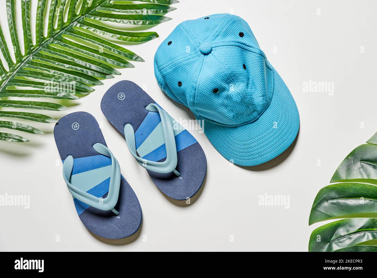 Zwei Paar blaue Flip Flops und ein Hut auf weißem Hintergrund mit Palmblättern im Vordergrund Stockfoto
