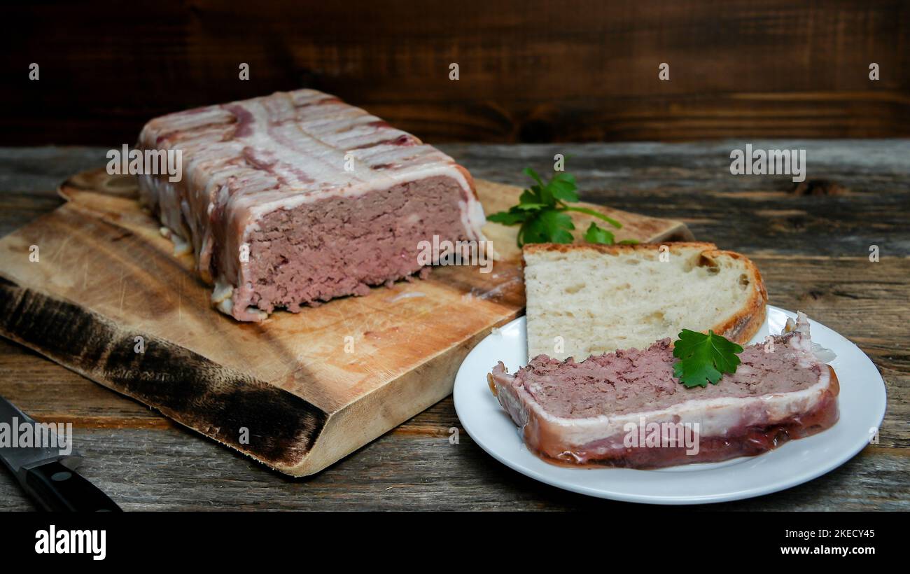 Hausgemachte Pastete, eingewickelt in Speck aus Schweinefleisch und Leber, auf einem rustikalen Tisch mit einer Scheibe auf dem Teller Stockfoto