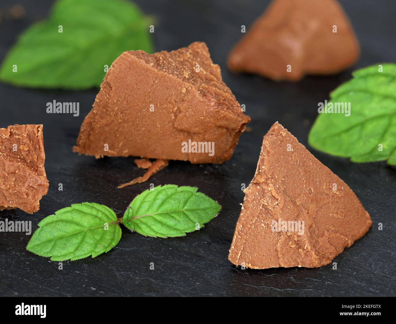 Süße Schokoladen-Haselnuss-Nougatstücke mit Minzblättern isoliert auf schwarzem Schieferbrett, als Backzutat oder süßer Aufstrich Stockfoto