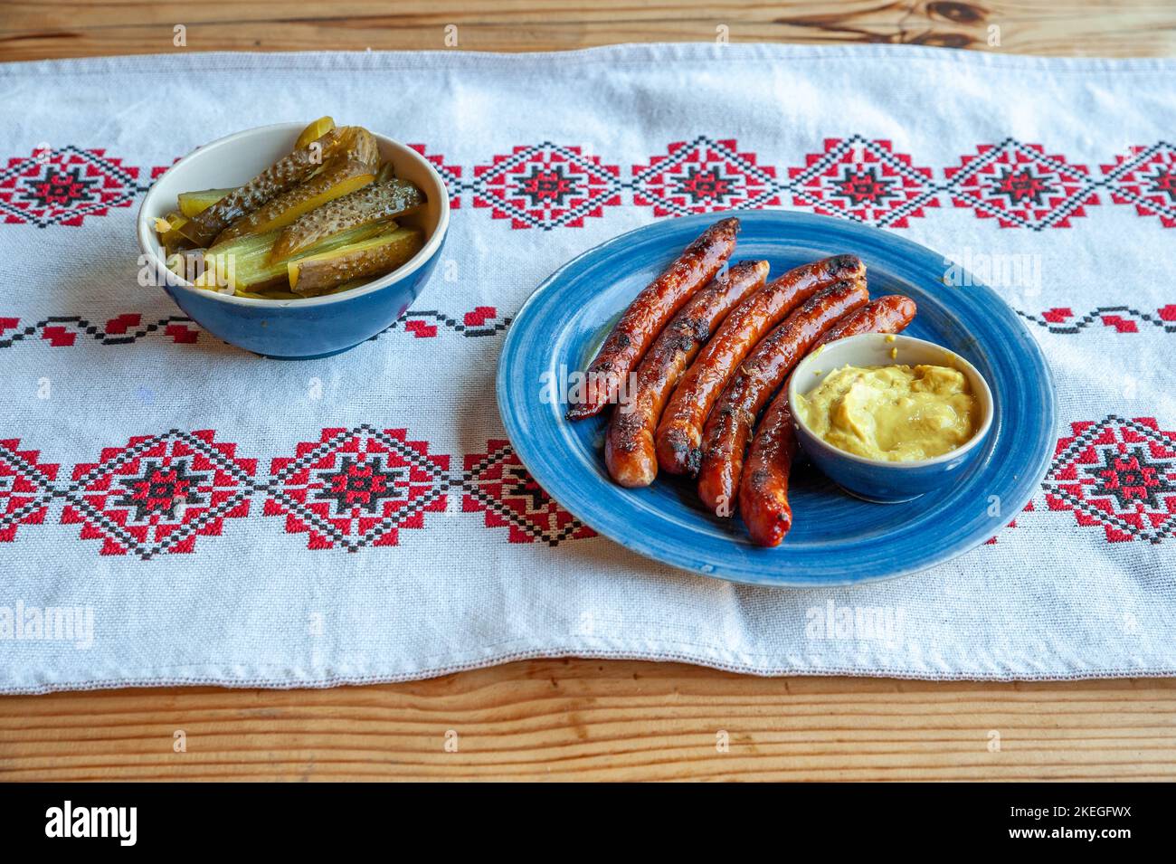 Eine schöne Aufnahme von gegrillten Würstchen, Kartoffelpüree, Gurken in Tongeschirr auf russische Stickerei Tischdecke gelegt Stockfoto