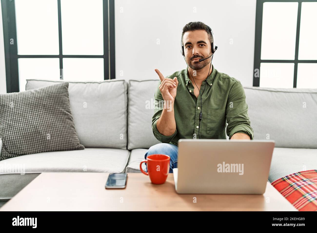 Junger hispanischer Mann mit Bart, der ein Callcenter-Agenten-Headset trägt und von zu Hause aus mit dem Finger zur Seite zeigt und Werbung zeigt, ernst Stockfoto