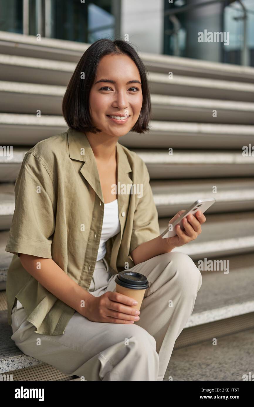 Vertikale Aufnahme des asiatischen Mädchens, das ihren Kaffee trinkt und das Mobiltelefon benutzt. Junge Frau mit Smartphone und Mitnahme sitzt auf Treppen Stockfoto