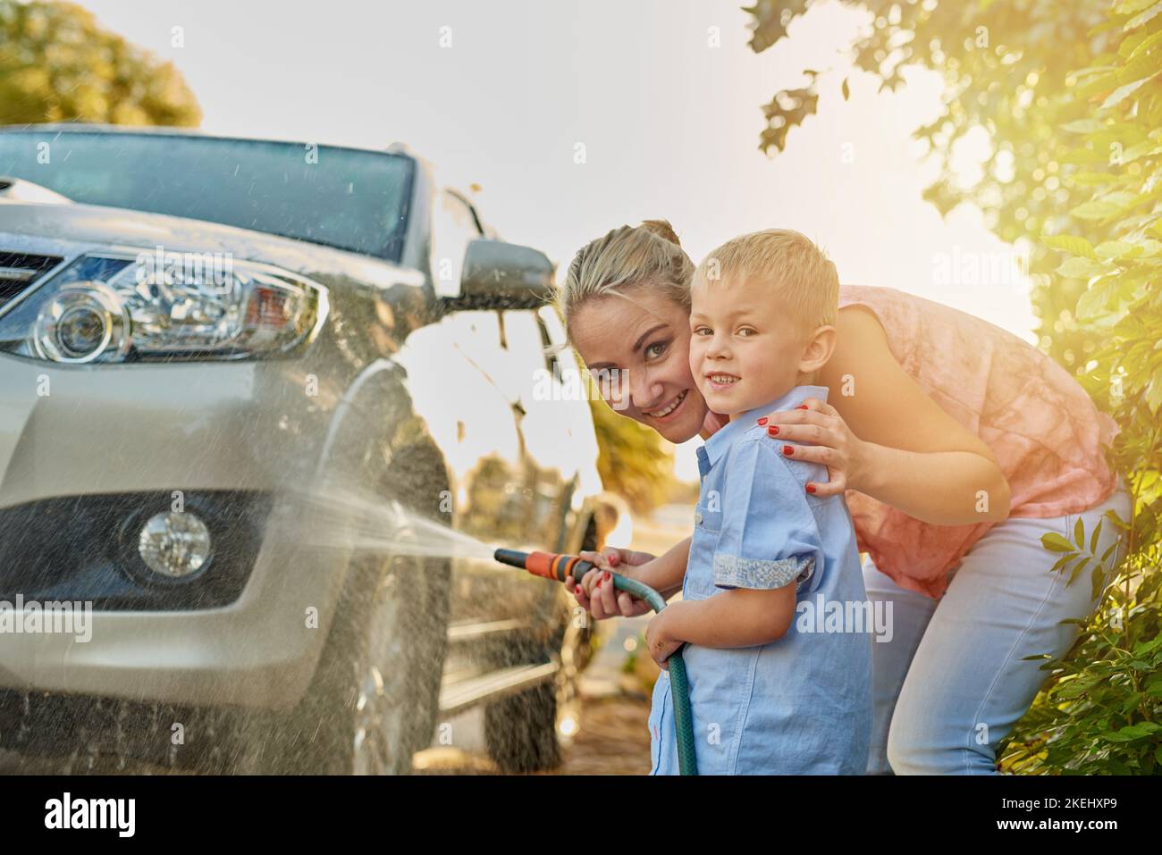 Das Auto zusammen mit dem Schlauchschaufeln abschmieren. Porträt einer Mutter und eines Sohnes, die zusammen ein Auto waschen. Stockfoto