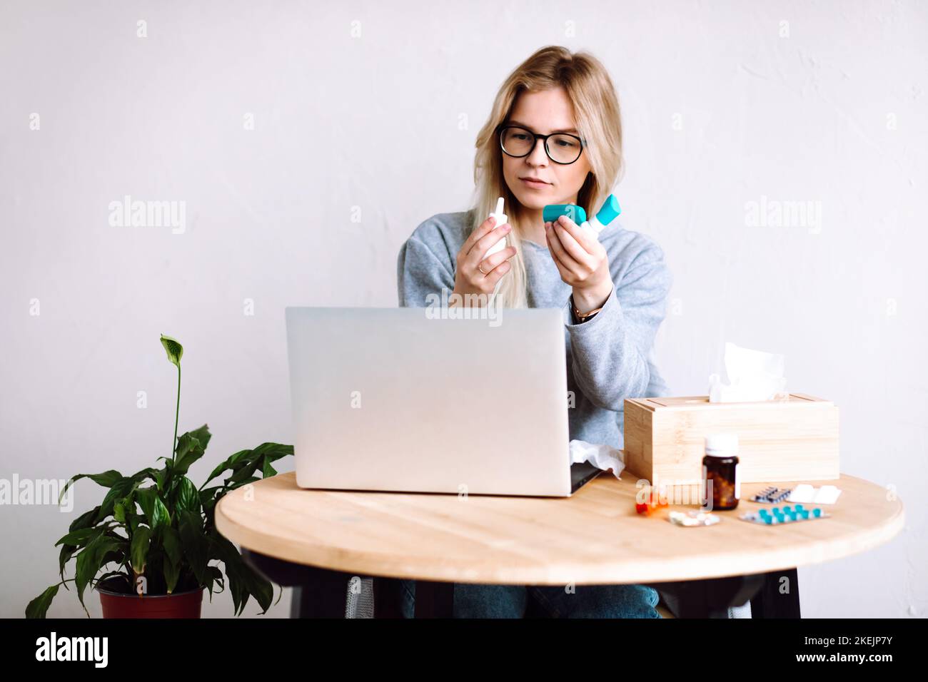 Porträt einer jungen Frau, die in der Nähe von Laptop-Box mit Papierservietten sitzt, mit Flaschen für Halsschmerzen, Nasensprayflasche. Stockfoto