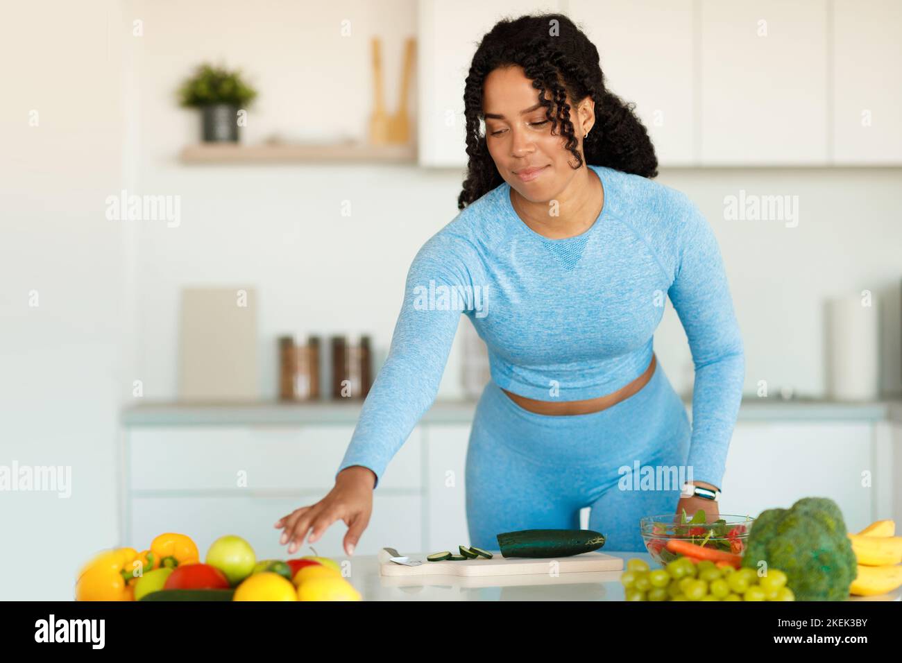 Junge afroamerikanische sportliche Dame schneidet Gurken und nimmt Paprika, bereitet frischen Gemüsesalat, kopieren Raum Stockfoto