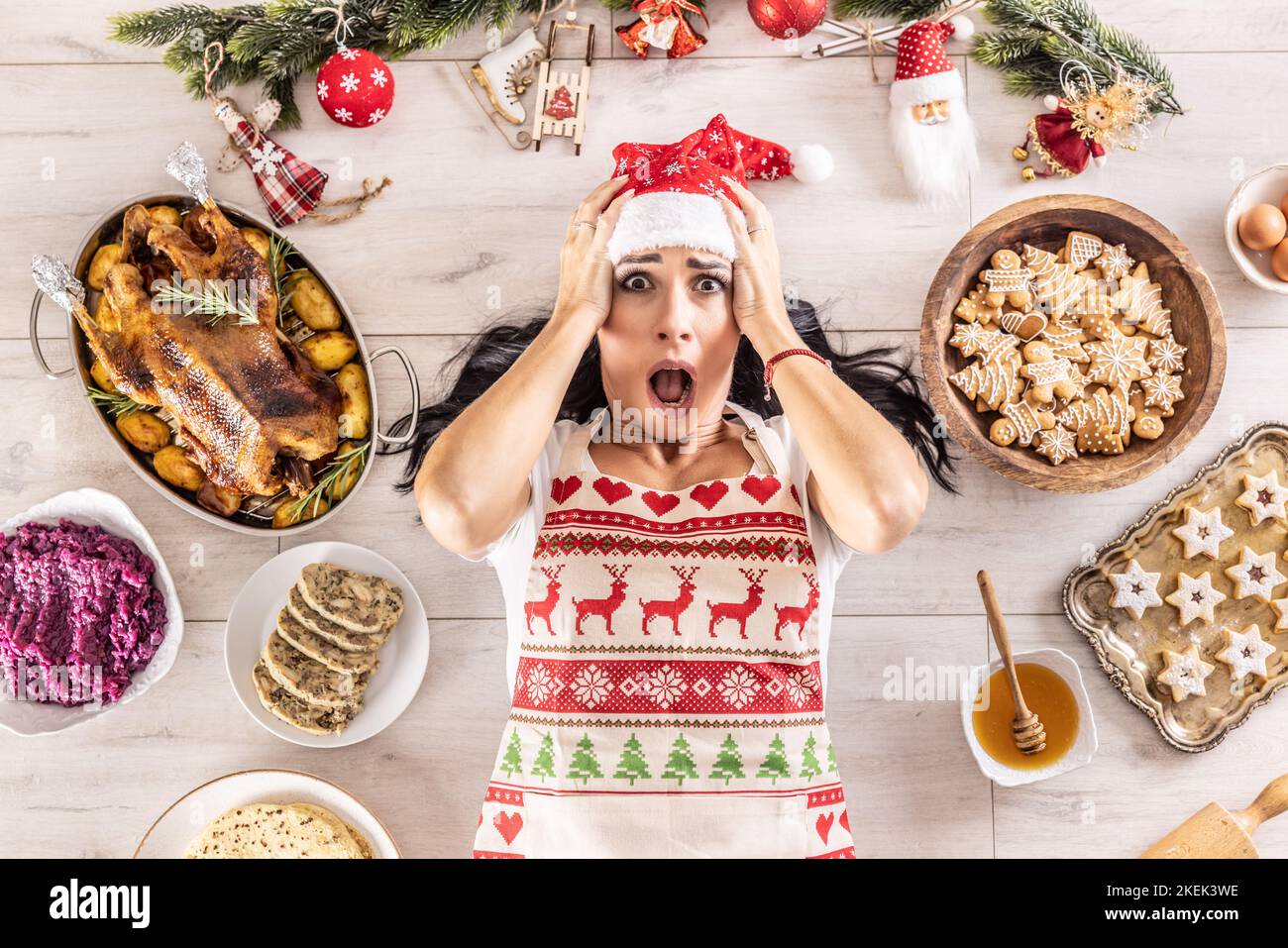 Gestresste Köchin in einer weihnachtlichen Schürze und Weihnachtsmütze, die ihren Kopf hält und auf dem Boden liegt, umgeben von Lebkuchen, Linzer Kuchen, auf der anderen Seite Stockfoto