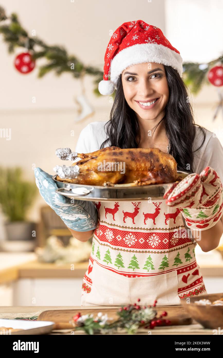 Zufriedene junge Frau in einer Weihnachtsschürze und Weihnachtsmütze mit einer Bratpfanne mit einer gebratenen Gans oder einem truthahn. Stockfoto