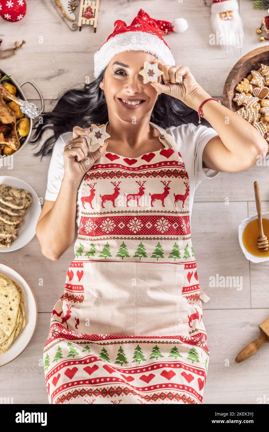 Eine fröhliche Köchin in einer weihnachtlichen Schürze liegt auf dem Boden und bedeckt ihre Augen mit Kuchen, umgeben von traditionellen Feiertagsgerichten und Kuchen. Stockfoto