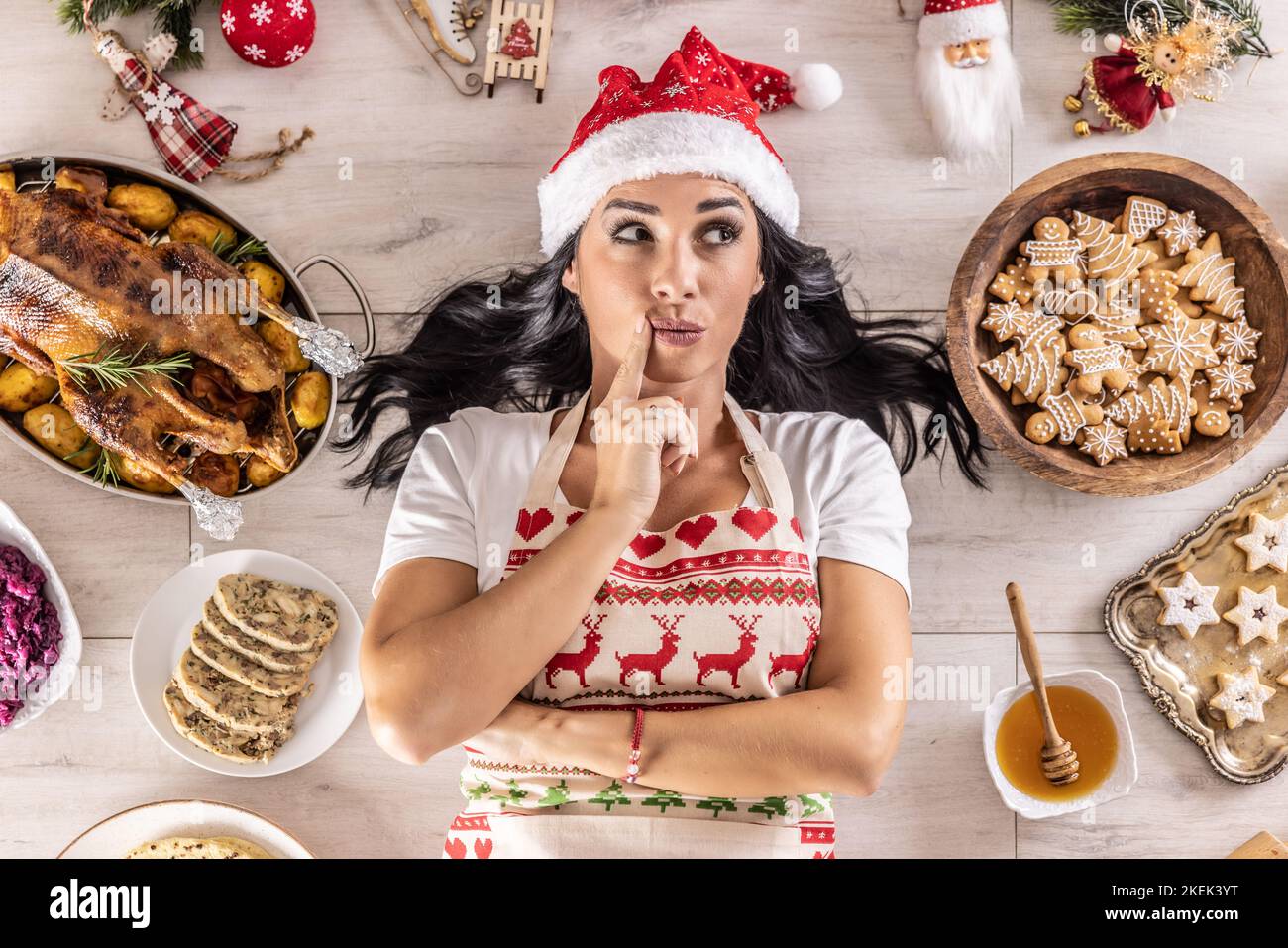 Nachdenkliche Köchin in Weihnachtsschürze und Weihnachtsmütze, auf dem Boden liegend, umgeben von Lebkuchen, Linzer Kuchen, auf der anderen Seite: Gebratene Gans oder tu Stockfoto