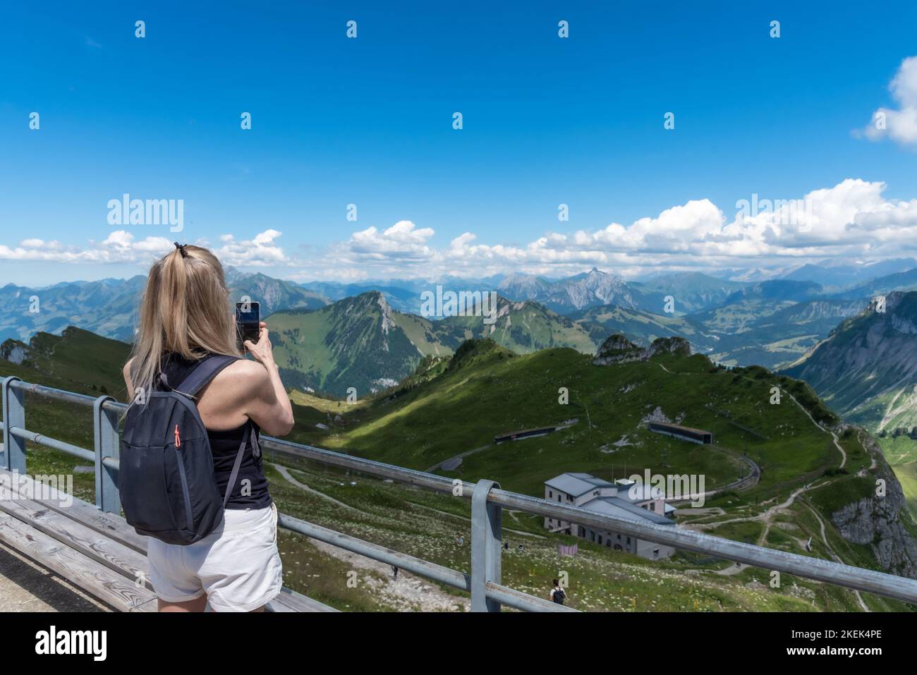 Reife Erwachsene blonde Frau mit Rucksack, die von einem Aussichtspunkt aus ein Foto der Schweizer Alpen fotografiert. Stockfoto