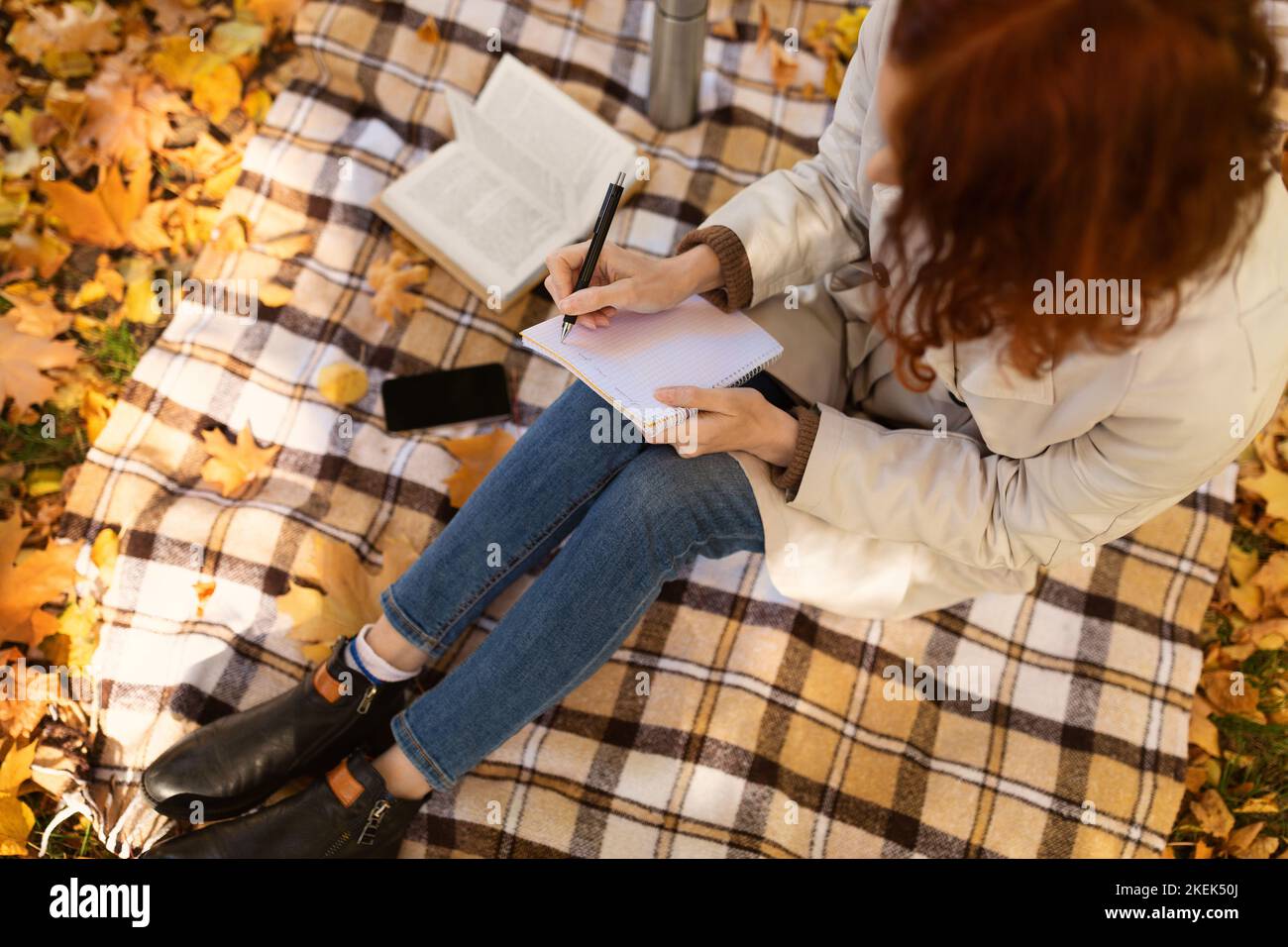 Kaukasische junge Studentin mit roten Haaren in Regenmantel sitzt auf karierten mit Telefon mit leerem Bildschirm Stockfoto
